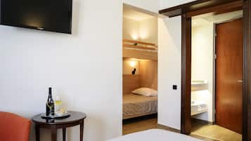 A hotel room with a bed, a small table with a bottle and glasses, a TV mounted on the wall, and a wooden door leading to a bathroom.