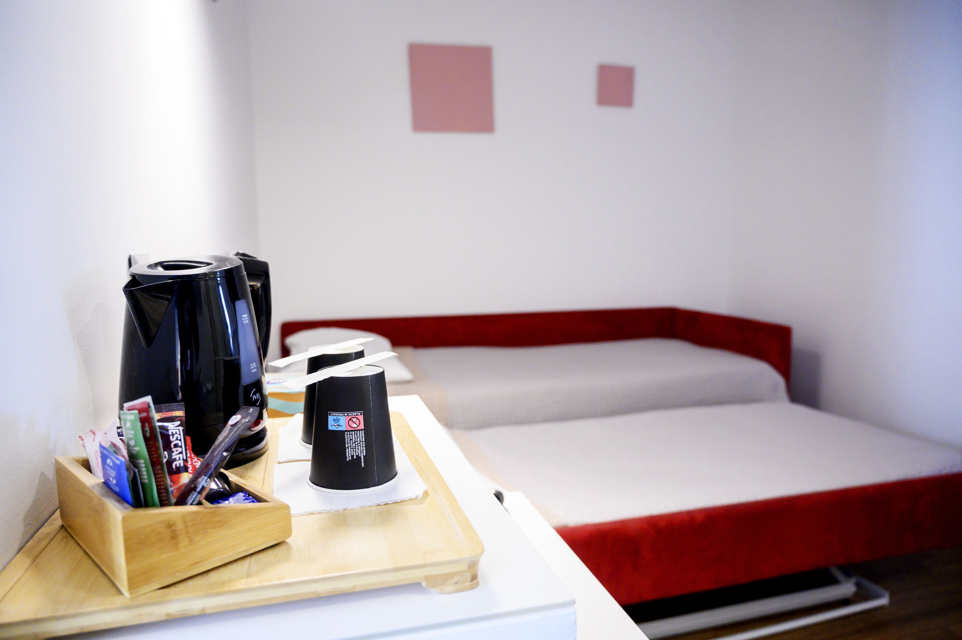 A hotel room with a bed, a small table with a coffee maker and a tray of snacks, and a white wall with two red square decorations.