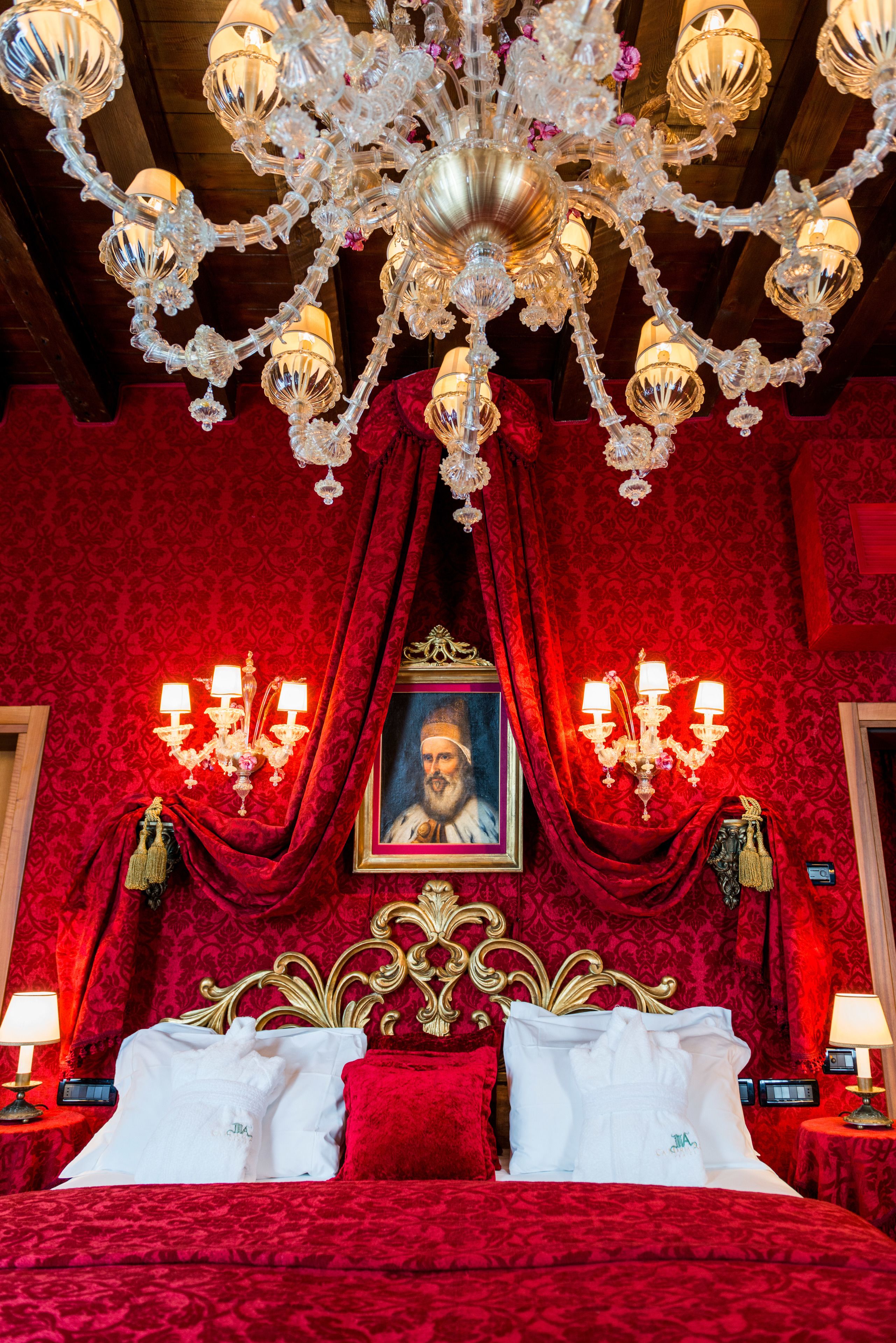 A luxurious room with a large bed, ornate chandelier, and a portrait on the wall.