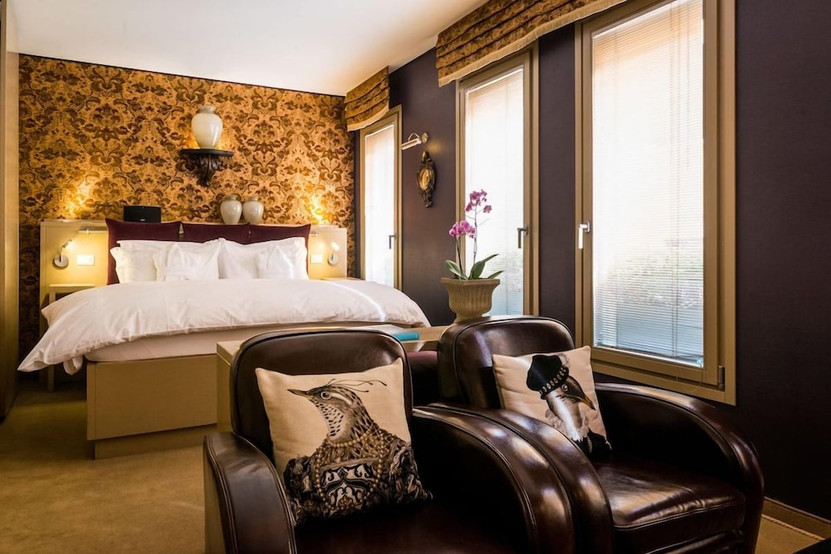 Een hotelkamer met een bed, een stoel met decoratieve kussens, een nachtkastje met een lamp en een raam met jaloezieën.