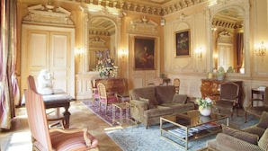 Een luxueuze kamer met versierd plafond, een grote bank, een salontafel en een open haard.