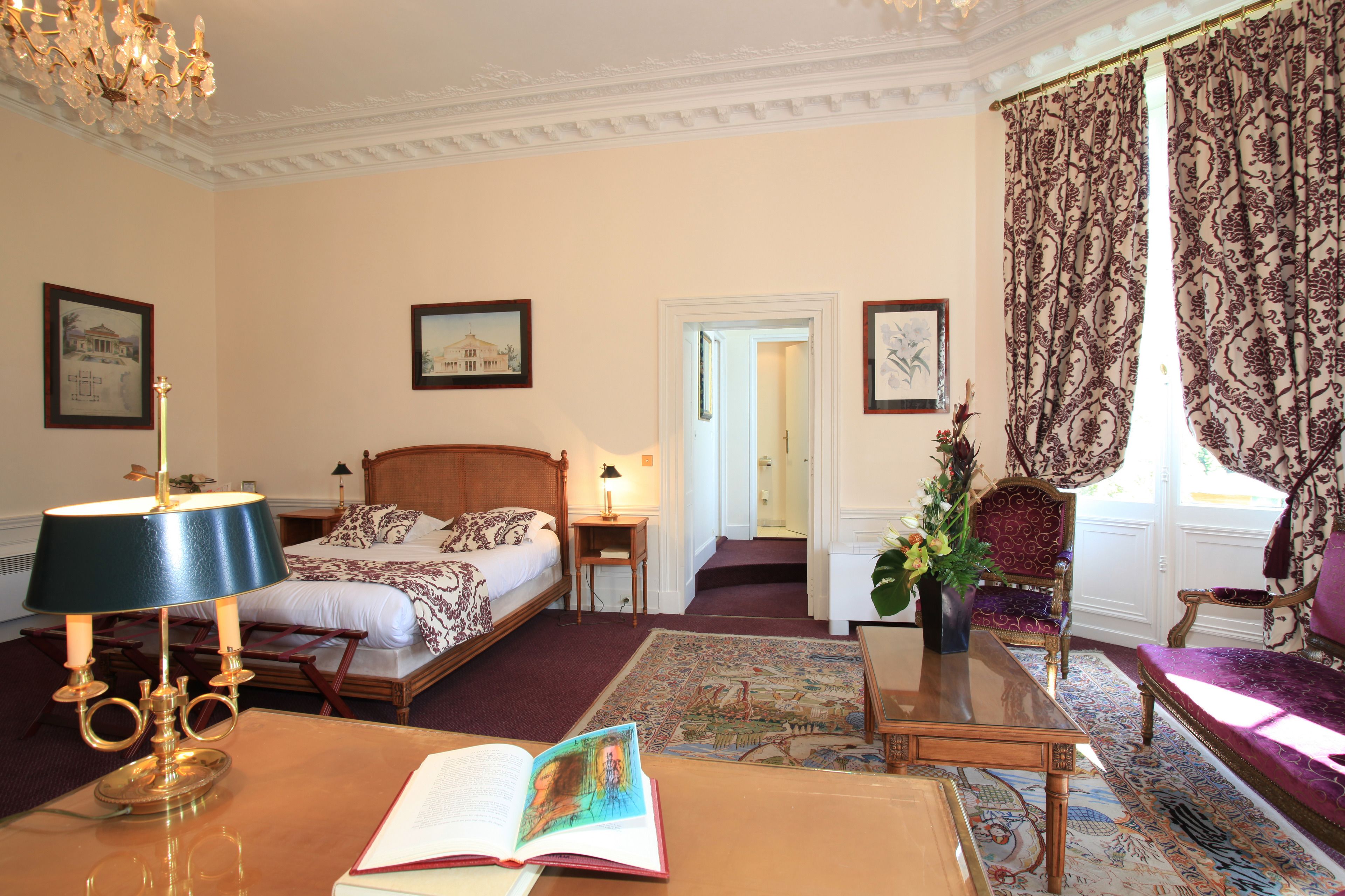 A hotel room with a bed, a desk with a lamp, a chair, a sofa, and a large window with curtains.