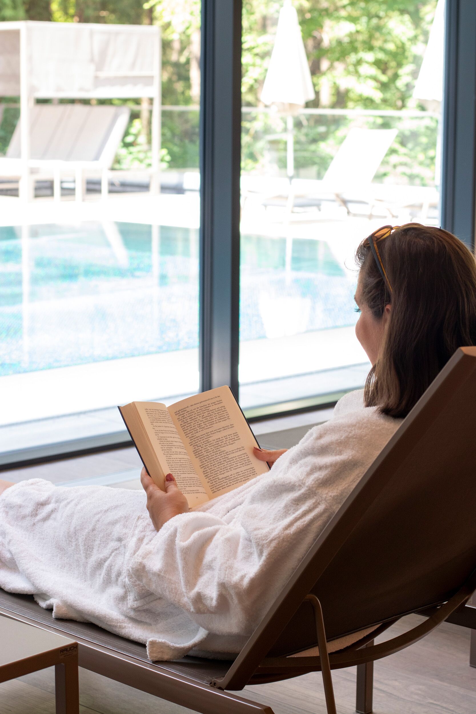 A person reading a book in a lounge chair by a poolside with a view of outdoor seating and umbrellas.
