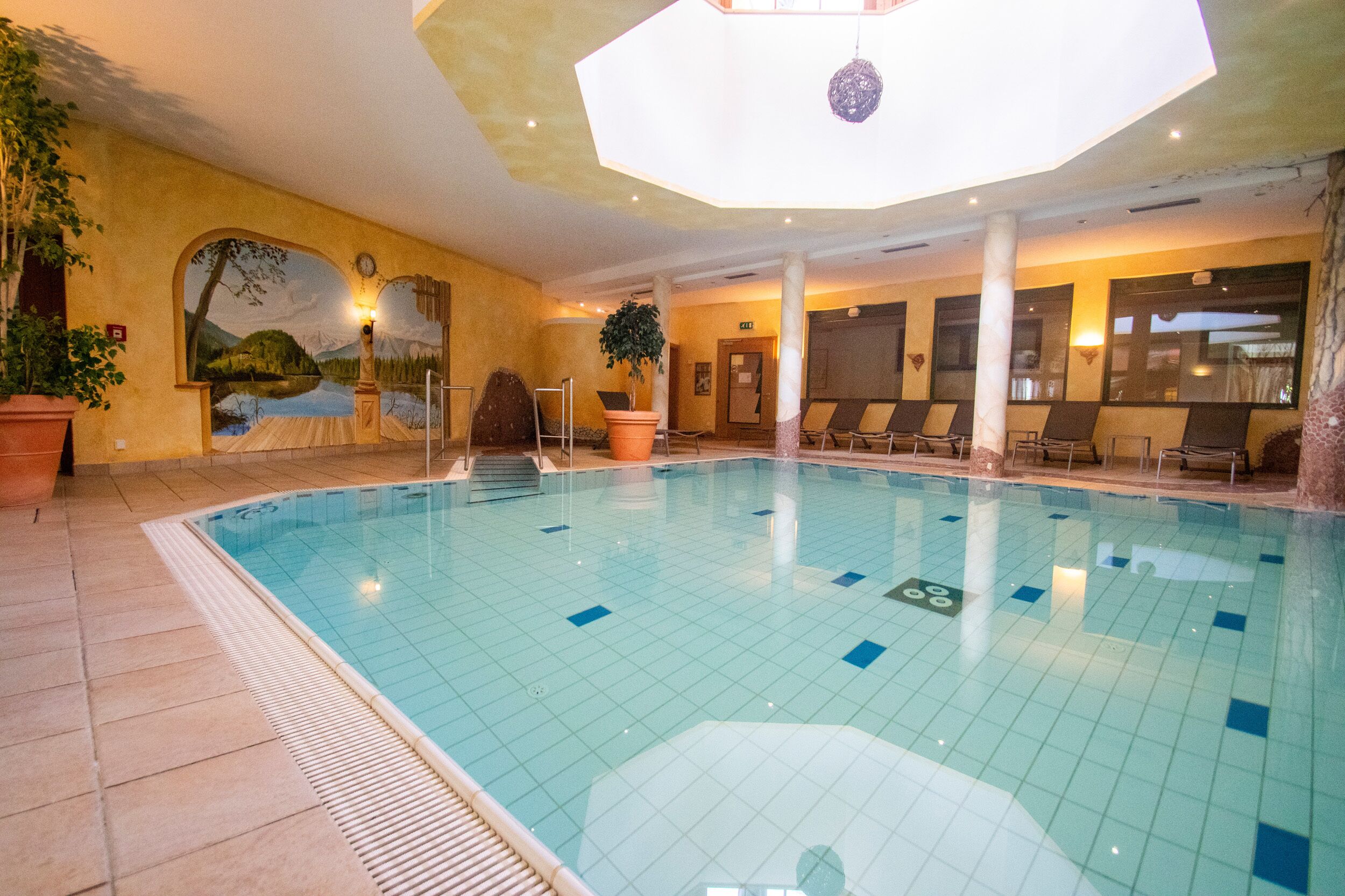 Ein Hallenbad mit gefliesten Wänden, Wandgemälden und Liegestühlen.