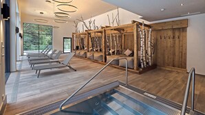 A modern indoor pool area with sun loungers, a wooden sauna, and a view of the outdoors. - Hotel Waldhof (Oetz)