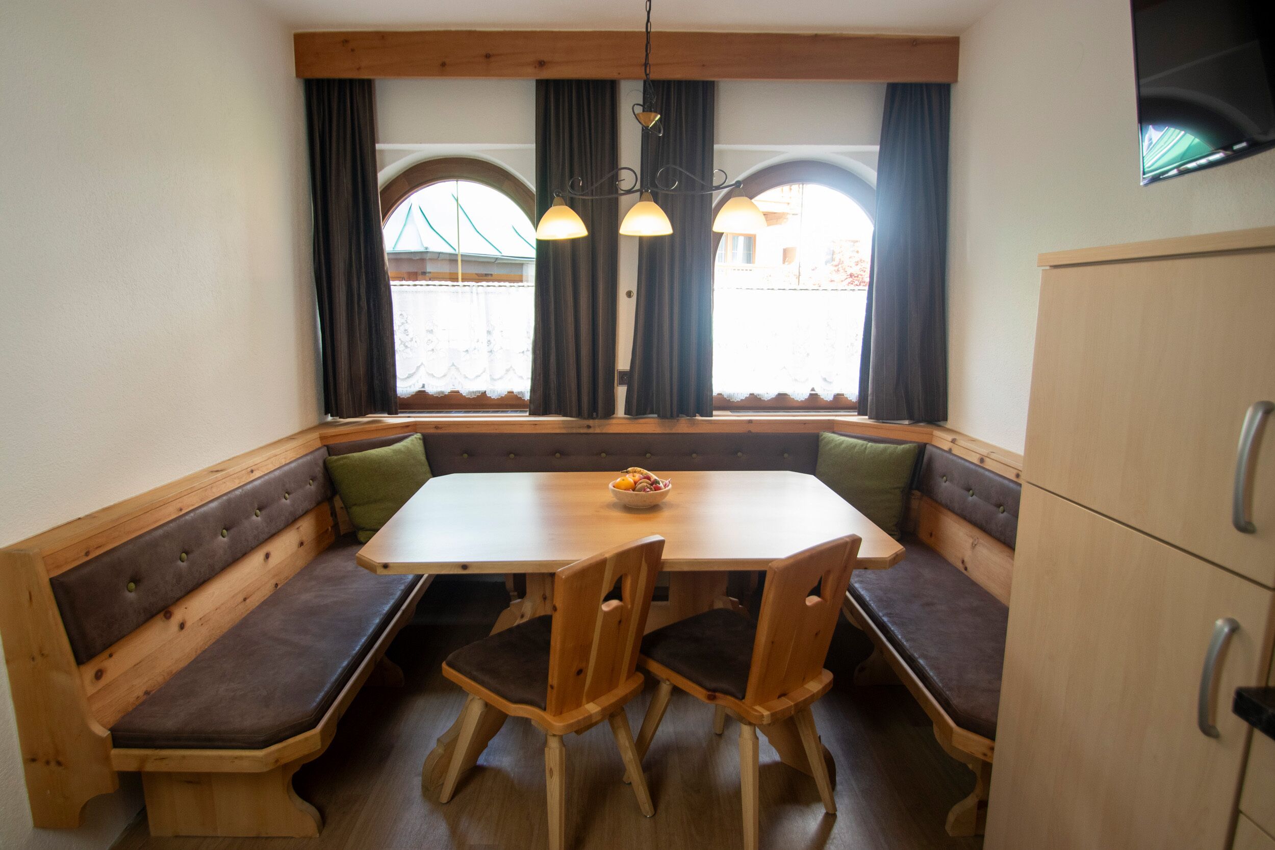 A dining area with a wooden table and bench seating, a bowl of fruit, and a television mounted on the wall.