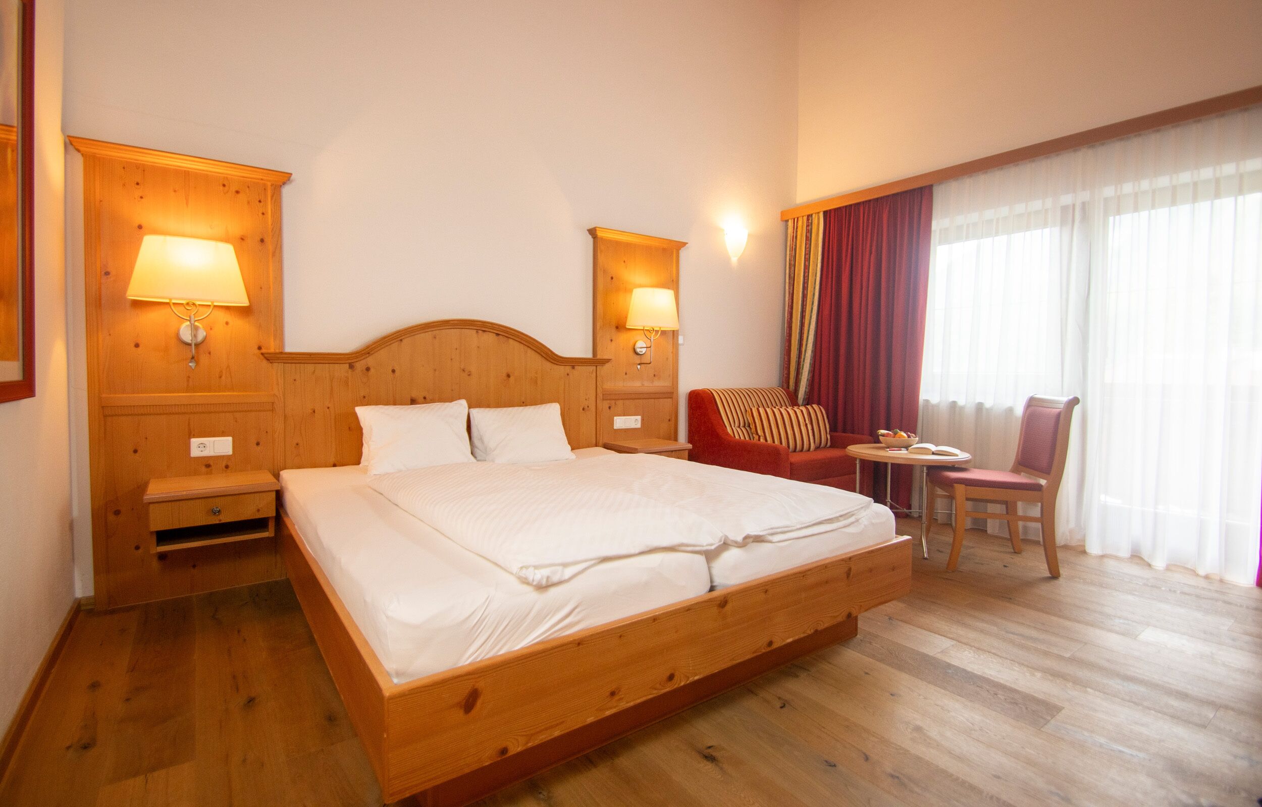 Quarto de hotel com cama de madeira, duas mesas de cabeceira com abajures, uma pequena mesa com cadeira e uma janela grande com cortinas.