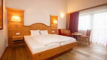 Quarto de hotel com cama de madeira, duas mesas de cabeceira com abajures, uma pequena mesa com cadeira e uma janela grande com cortinas.