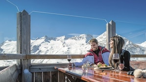 A wooden deck with a table set for two, overlooking a snowy mountain range. - Hotel Waldhof (Oetz)