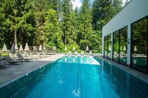 A swimming pool with lounge chairs and umbrellas, surrounded by trees and a modern building with large windows. - Hotel Waldhof (Oetz)