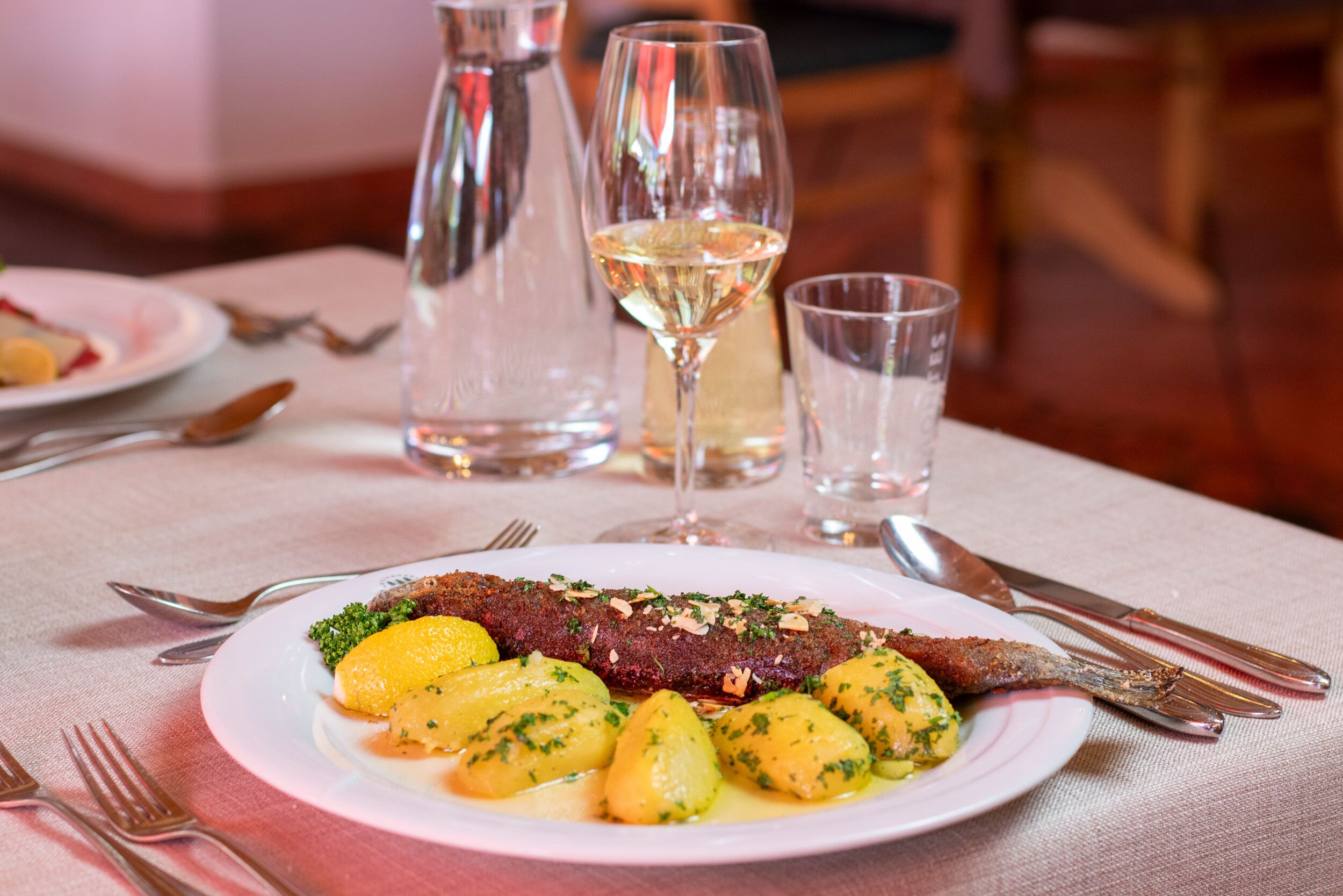 En tallrik med mat bestående av fisk, potatis och en citronskiva, placerad på ett bord tillsammans med bestick och glas.