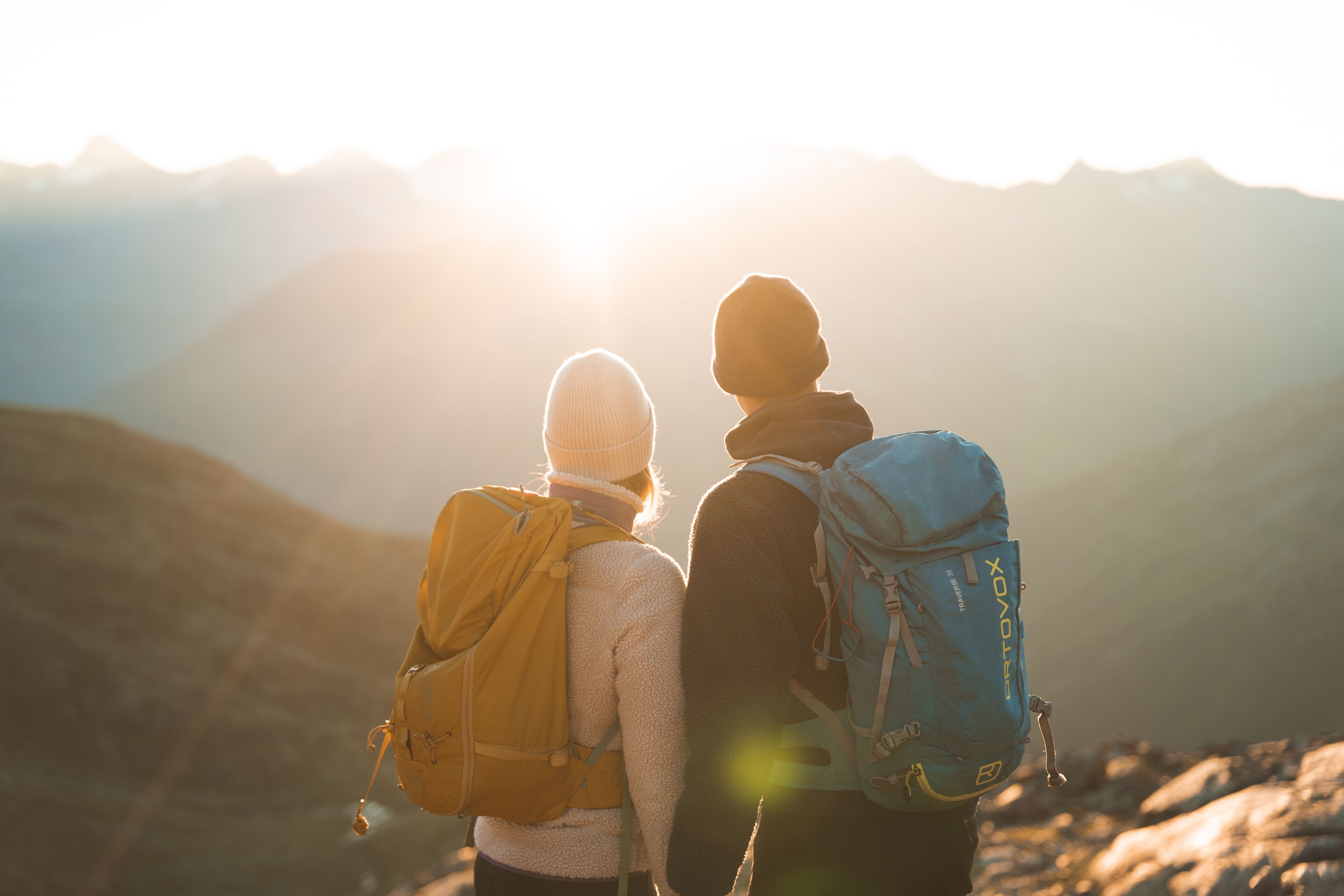 Dos personas con mochilas de espaldas a una cordillera, con el sol poniéndose al fondo.
