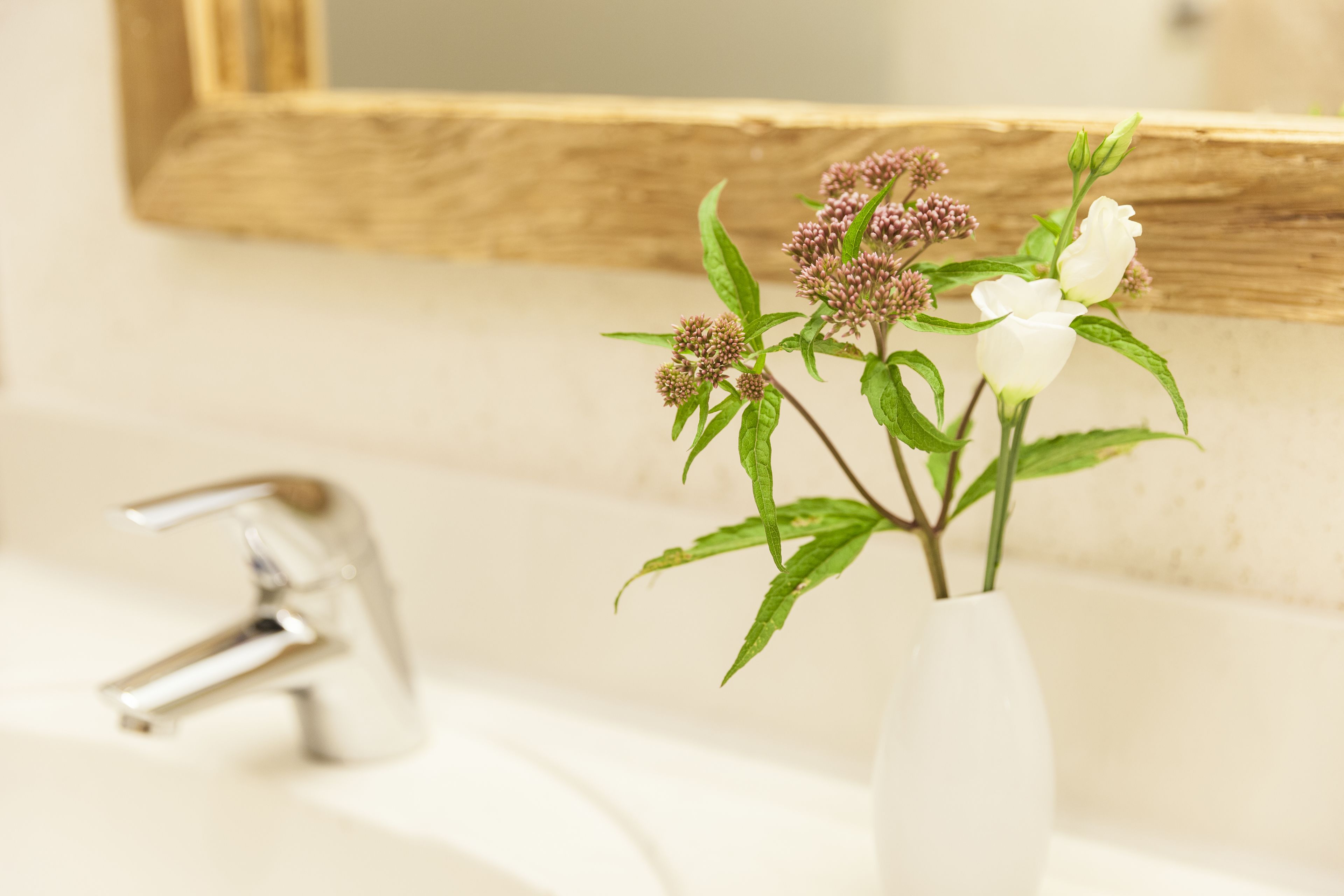 Un baño con un lavamanos blanco, un espejo con marco de madera y un florero con flores.