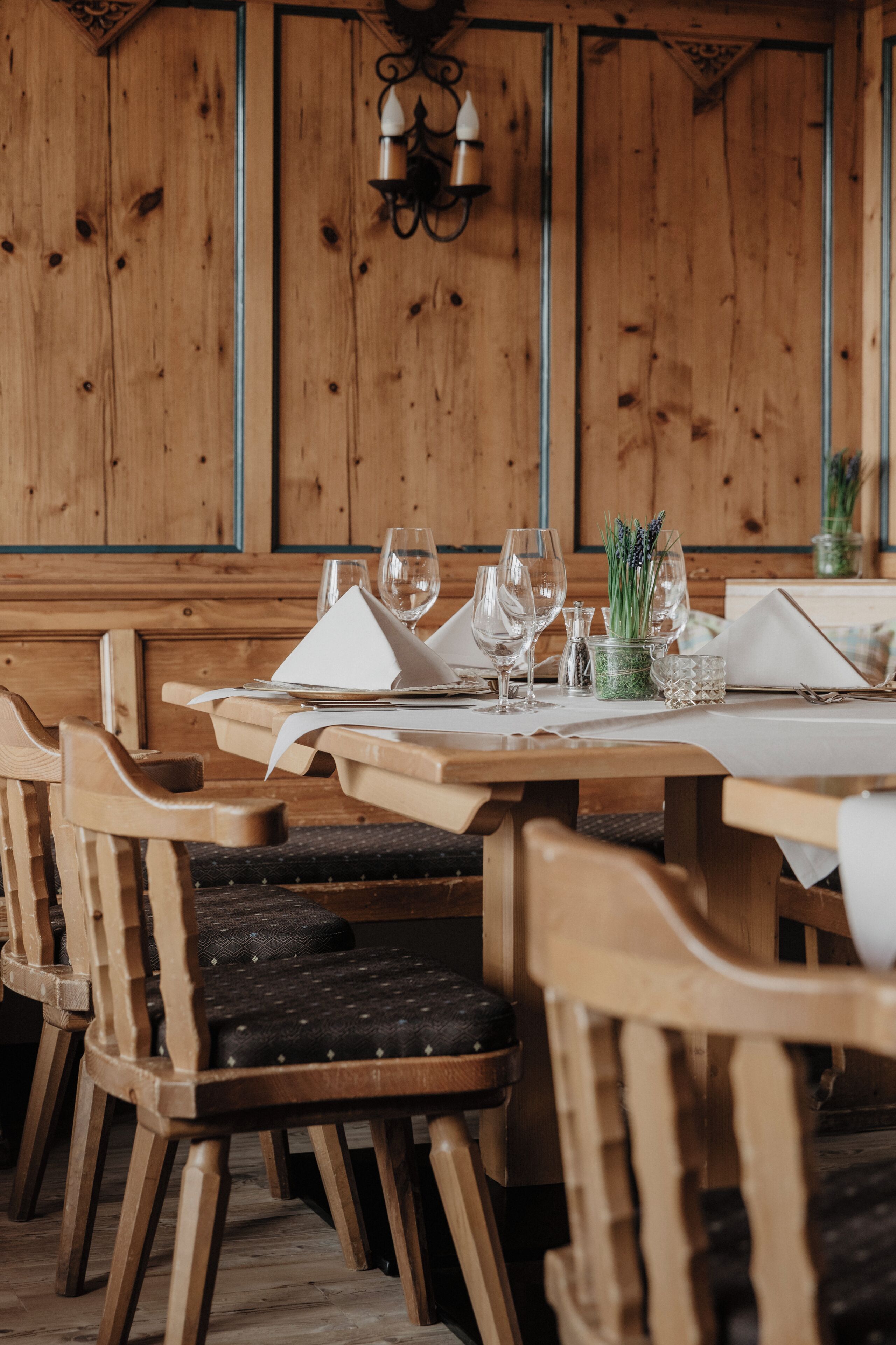 Een houten eettafel gedekt met witte servetten, wijnglazen en een kleine potplant.