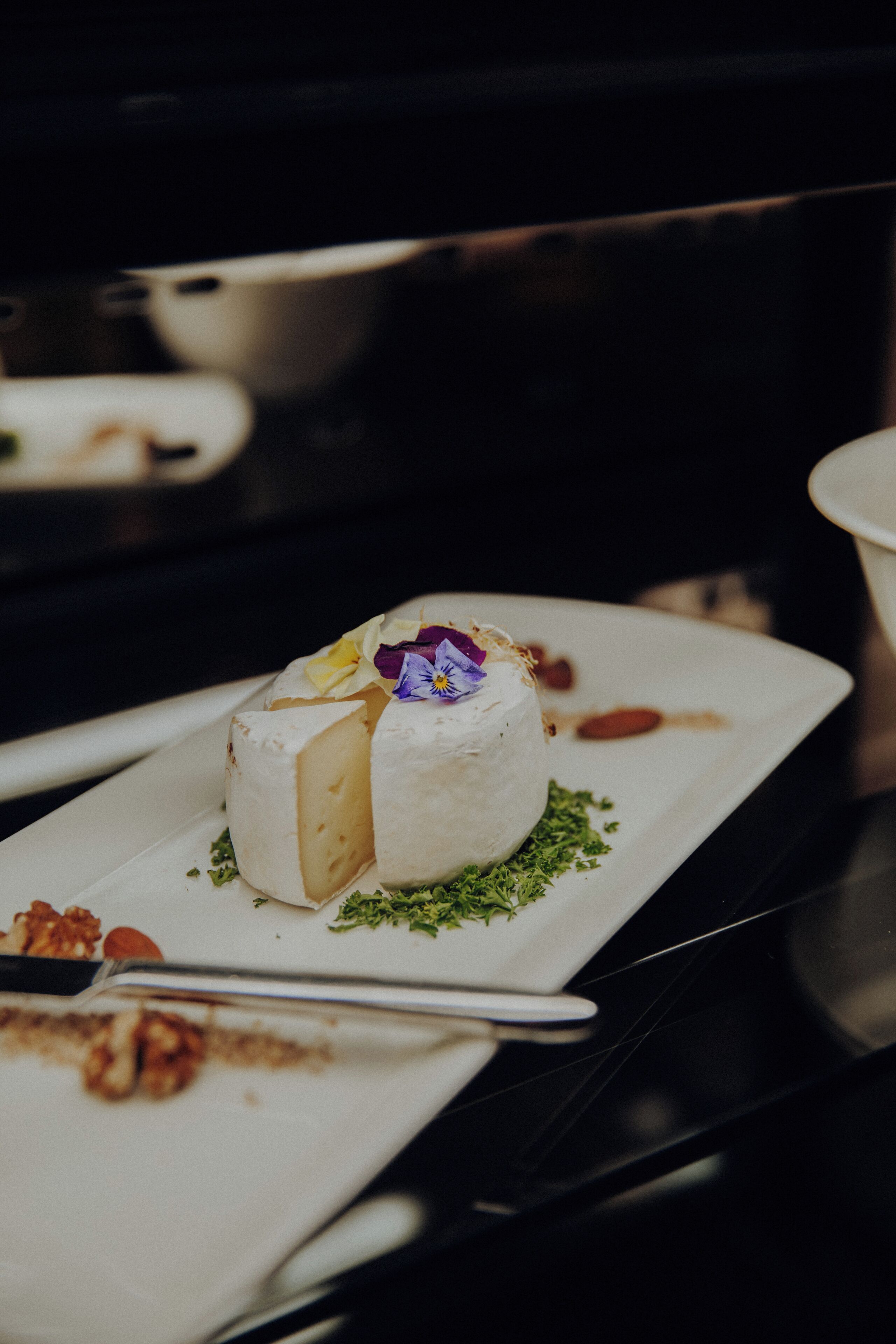 A white plate with a round cheese centerpiece, garnished with herbs and edible flowers, on a dark surface.