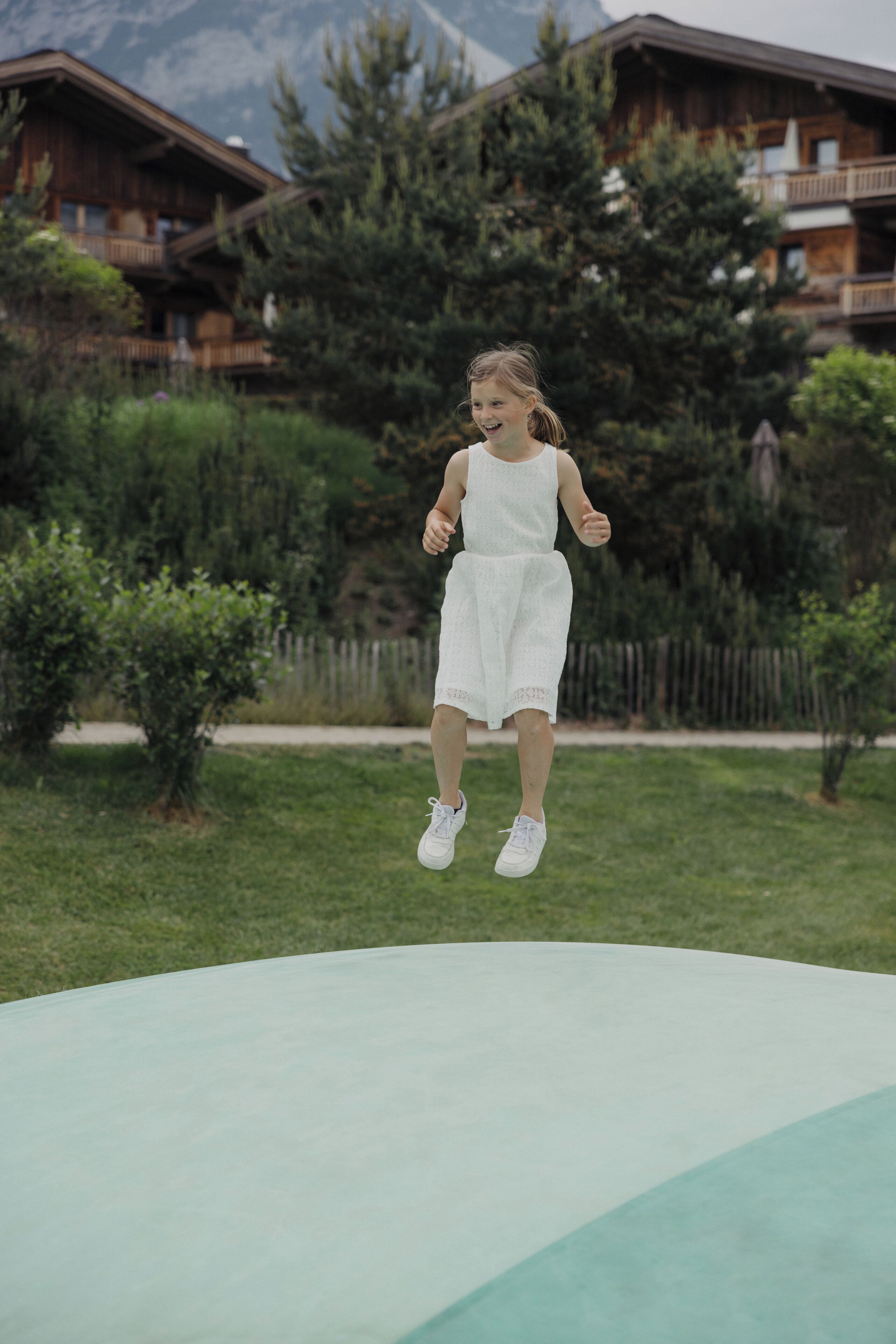 Eine Person springt auf einem Trampolin in einer grünen Umgebung, im Hintergrund sind Holzhütten zu sehen.