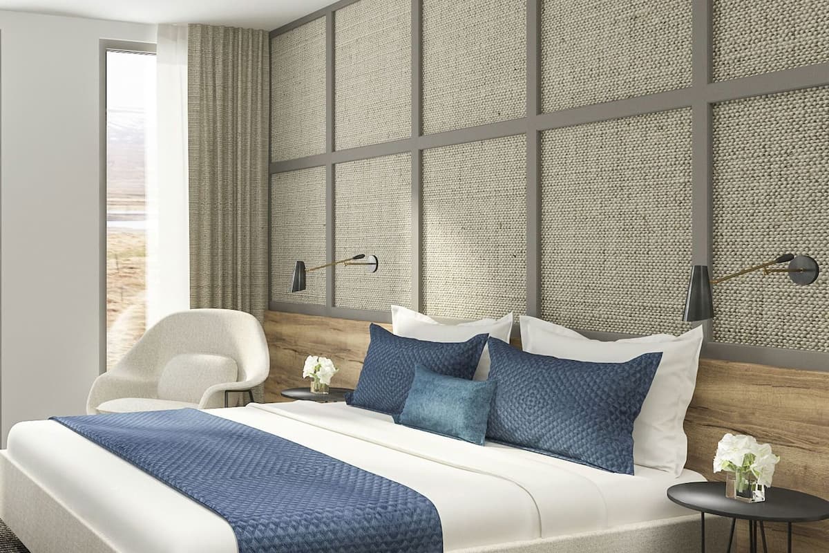 A modern hotel room with a large bed, a desk, a chair, and a view of a desert landscape through the glass door.