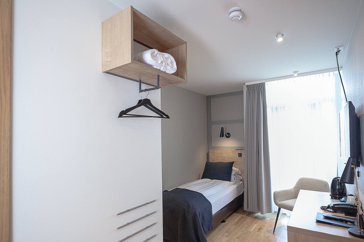 A modern hotel room with a bed, a desk, a hanging coat rack, and a ceiling-mounted light fixture.