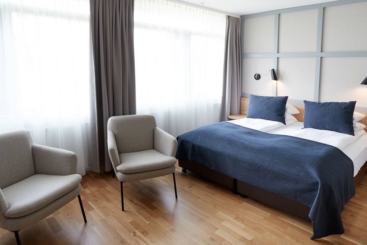 A hotel room with a bed, two armchairs, a large window with curtains, a wall-mounted lamp, and a wooden floor.