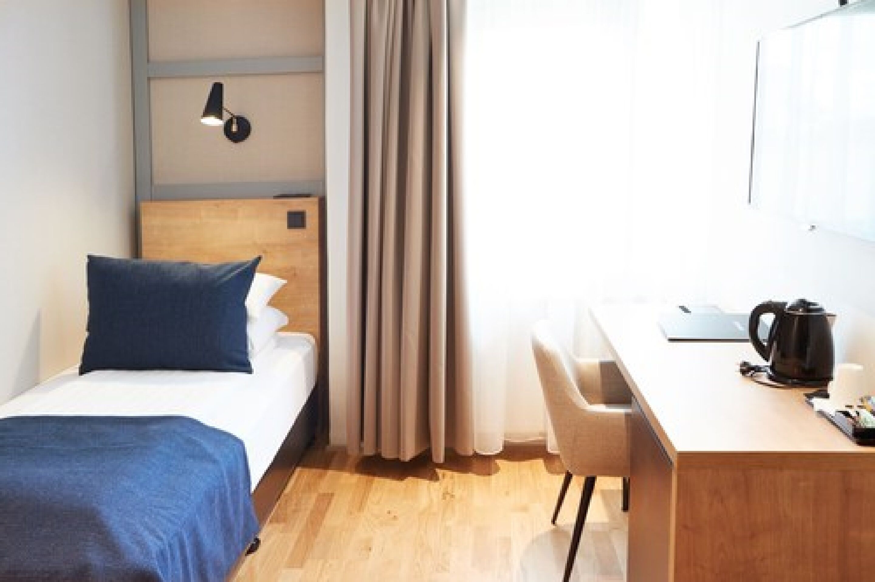 A hotel room with a bed, a desk, a chair, a lamp, and a curtain.