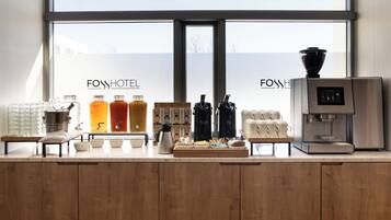 Zona de recepción de hotel con máquina de café, estación de té y varios dispensadores de bebidas.