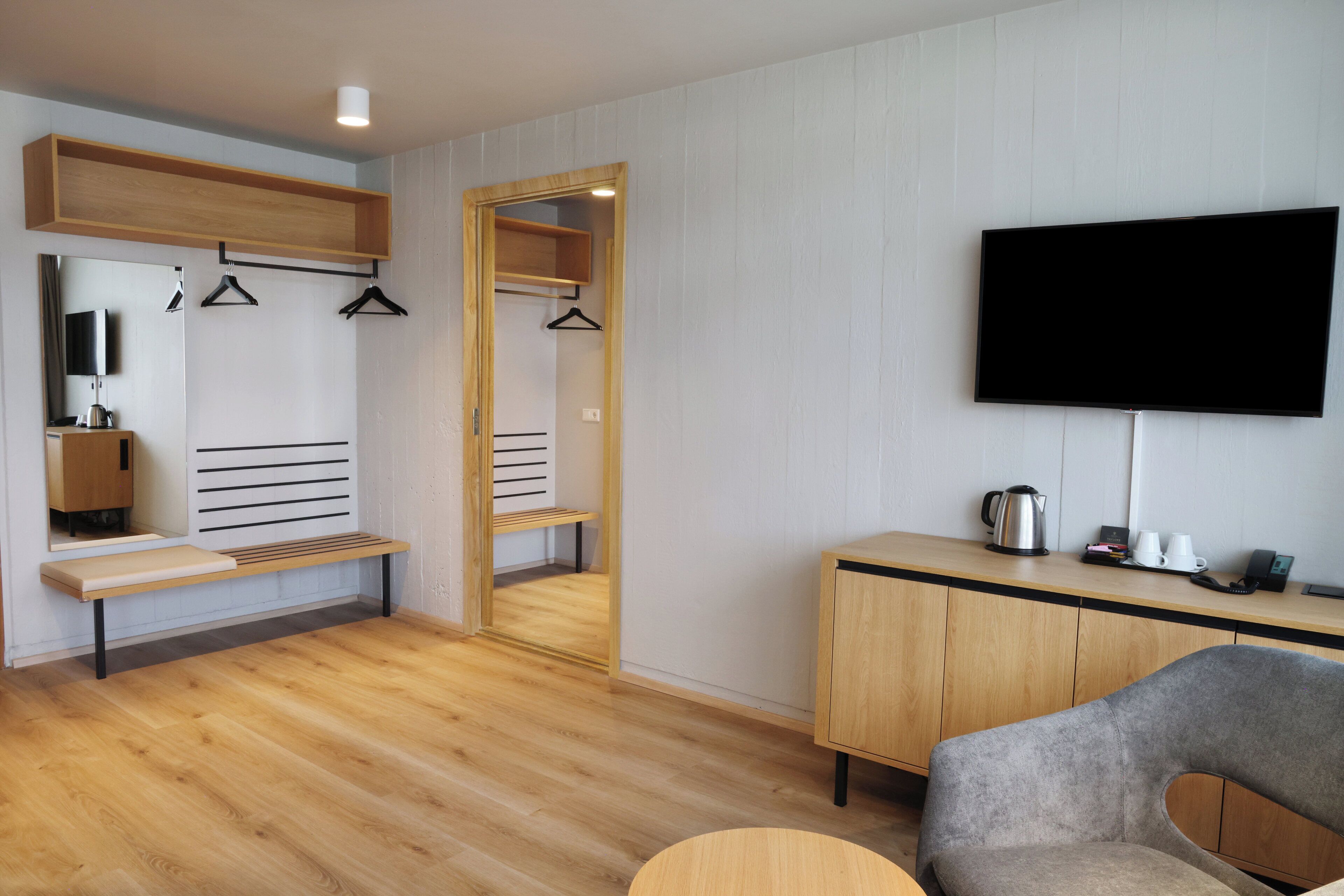 Ein modernes Zimmer mit einer Holzbank, einem an der Wand montierten Fernseher, einem Spiegel und einem Schrank mit Wasserkocher und Tassen.