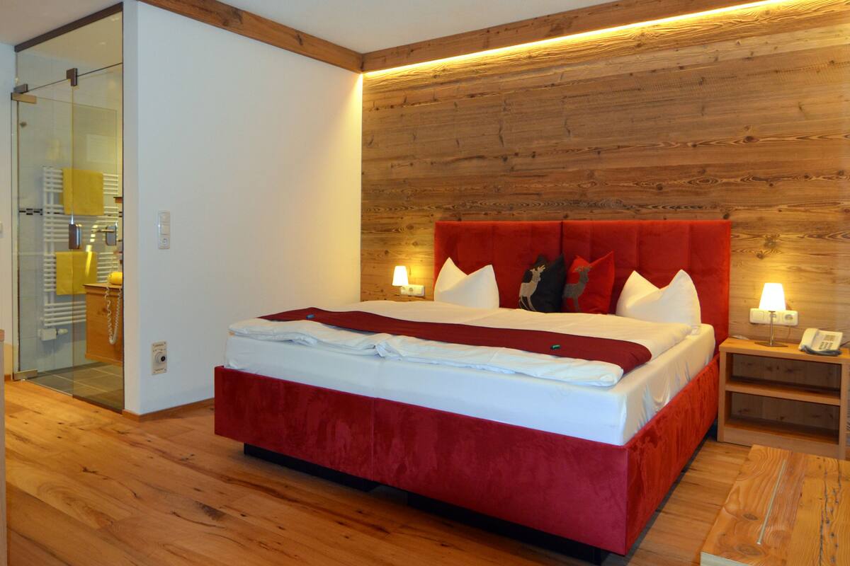 A modern hotel room with a wooden headboard, a large bed with red bedding, a bedside table with a lamp, and a glass shower enclosure.