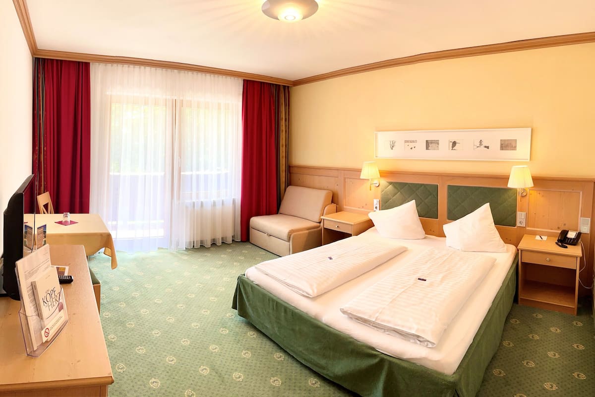 A hotel room with a bed, two bedside tables, a desk, a chair, a sofa, and a window with curtains.