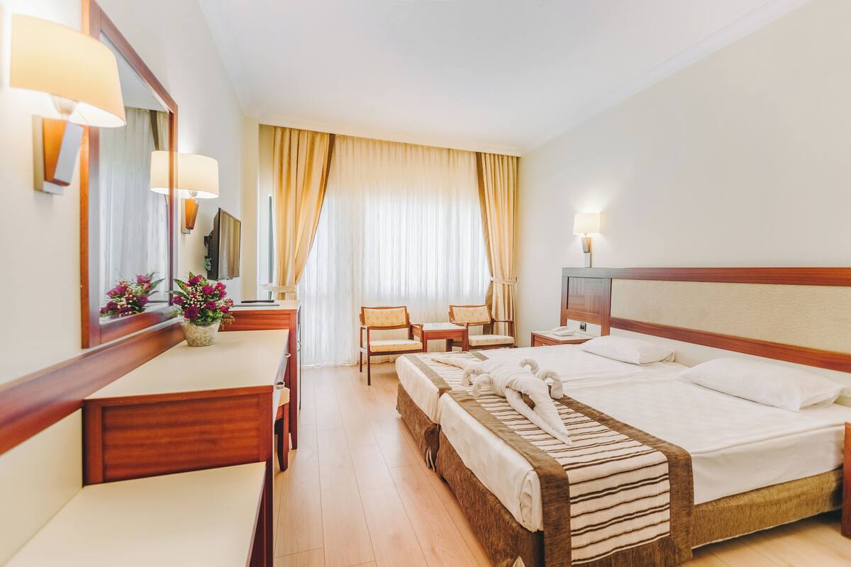 Una habitación de hotel con cama, escritorio, dos sillas, un espejo y una ventana con cortinas.