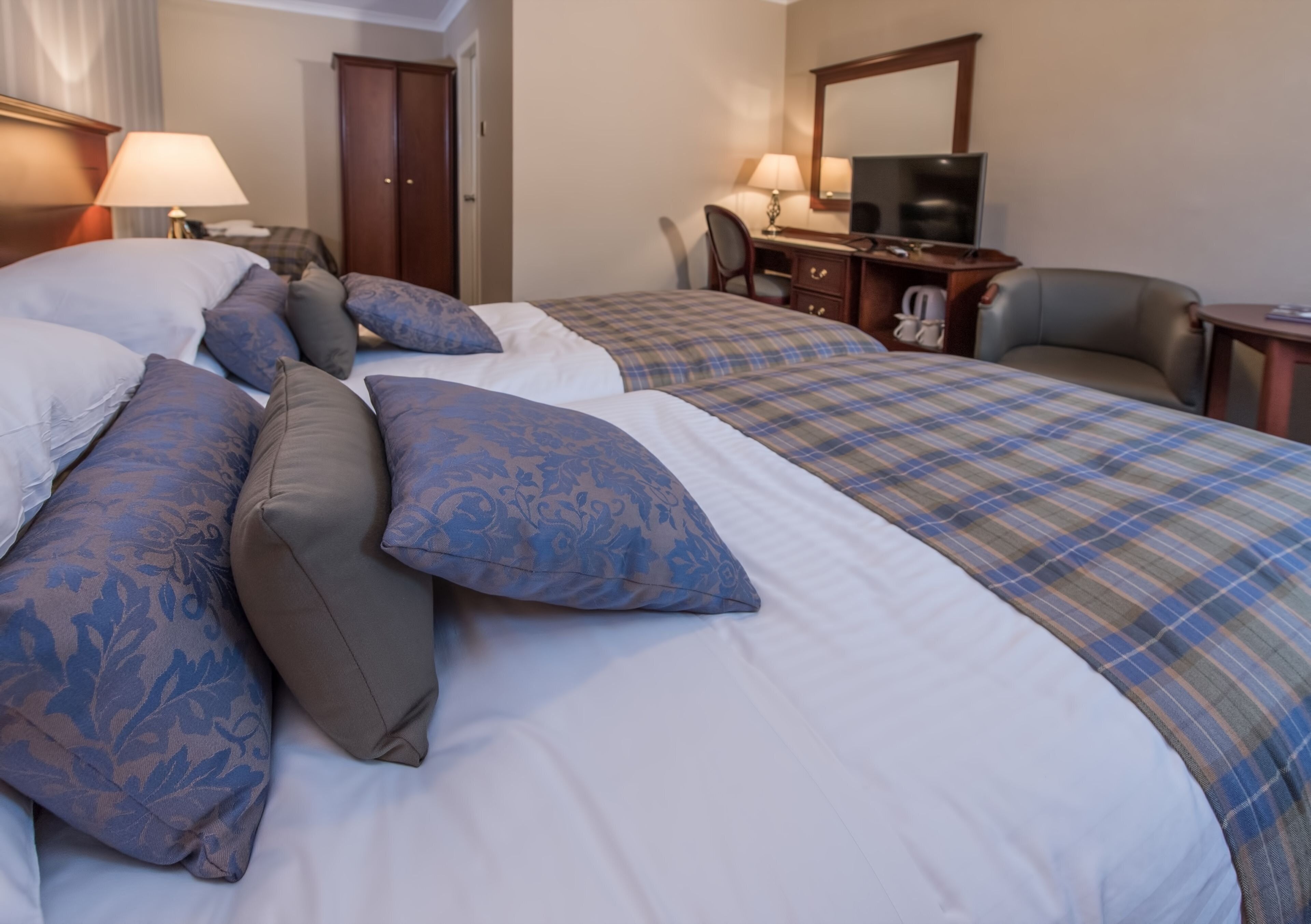 A hotel room with two beds, each with a different style of bedding. There is a desk with a chair, a television, and a telephone.