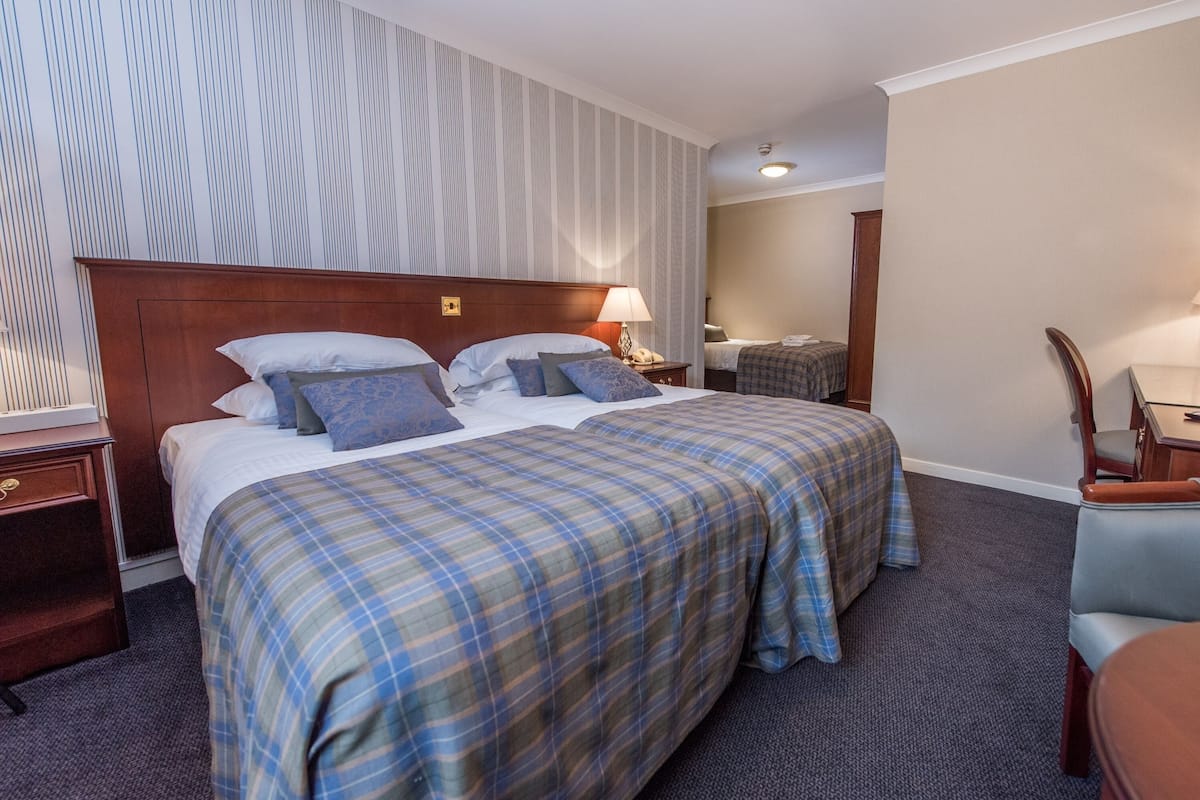 A hotel room with two beds, a wooden headboard, a nightstand with a lamp, a chair, and a small table.