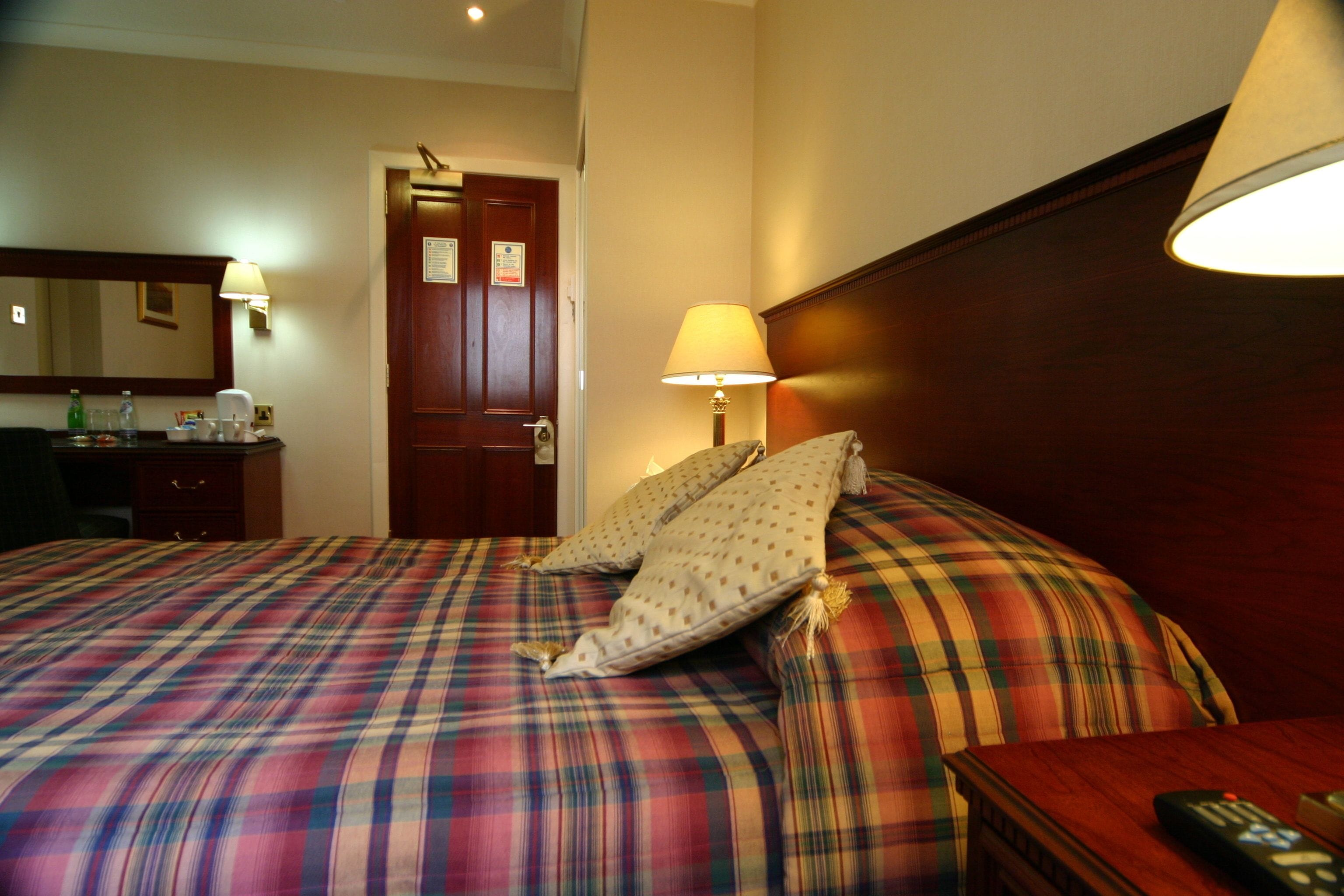 A hotel room with a plaid bedspread, a wooden headboard, a bedside table with a lamp, and a door with a sign.