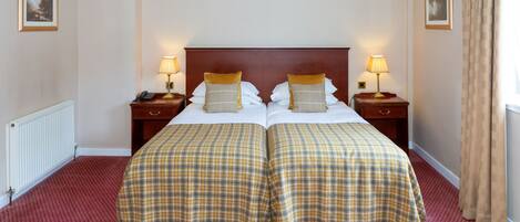 A double bed with a plaid bedspread and two pillows, flanked by nightstands with lamps, in a room with a red carpet and beige walls.