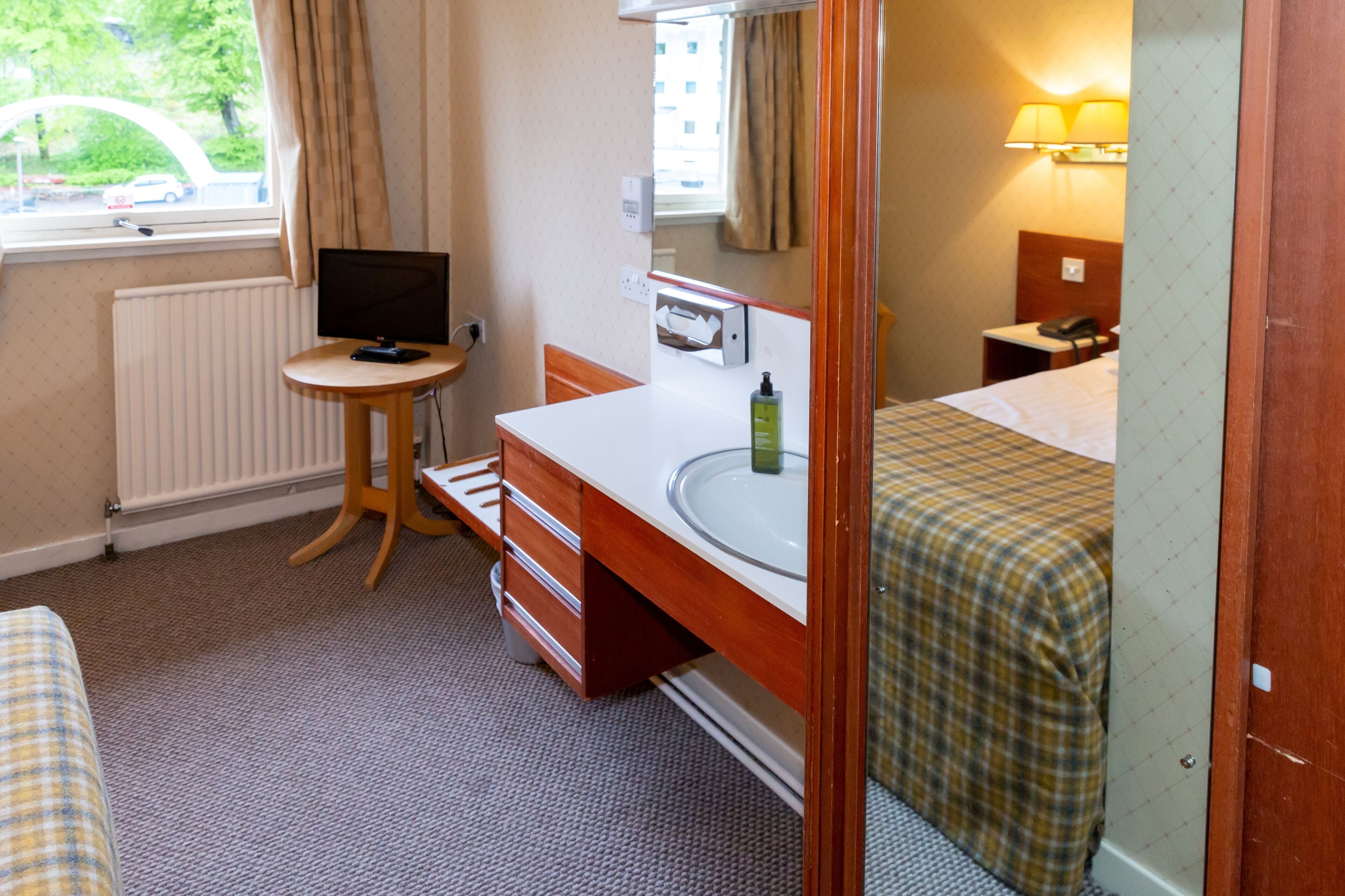 A hotel room with a bed, a bedside table with a lamp, a small desk with a chair, a window with curtains, and a radiator.