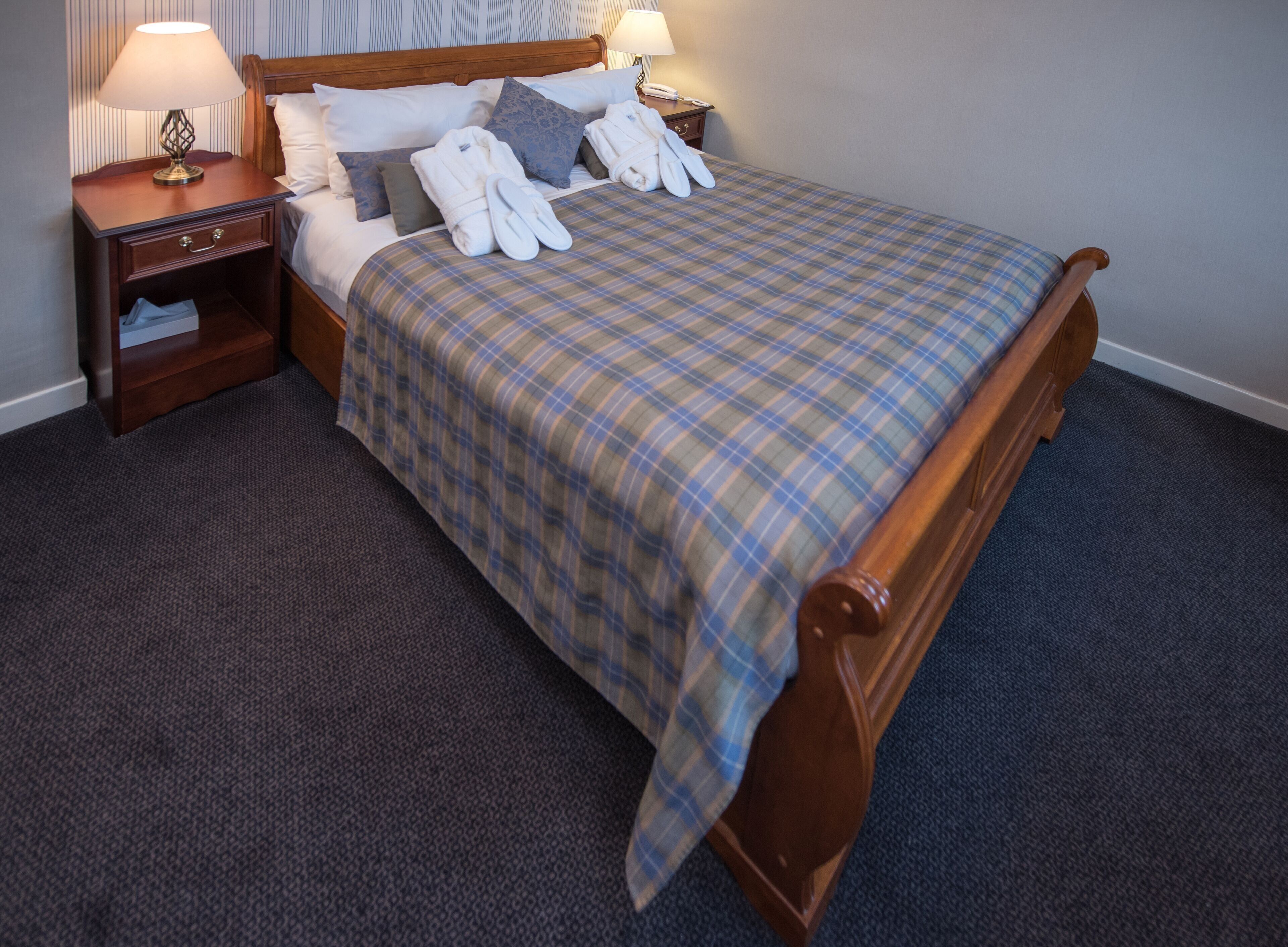 A neatly made bed with a plaid bedspread, two pillows, and a pair of white bathrobes. There are two bedside tables with lamps, one of which has a telephone. The room has a carpeted floor and wooden headboard.