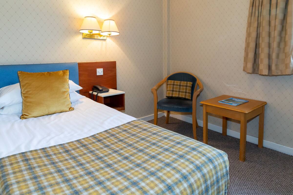 A hotel room with a bed, a chair, a small table, and a lamp.