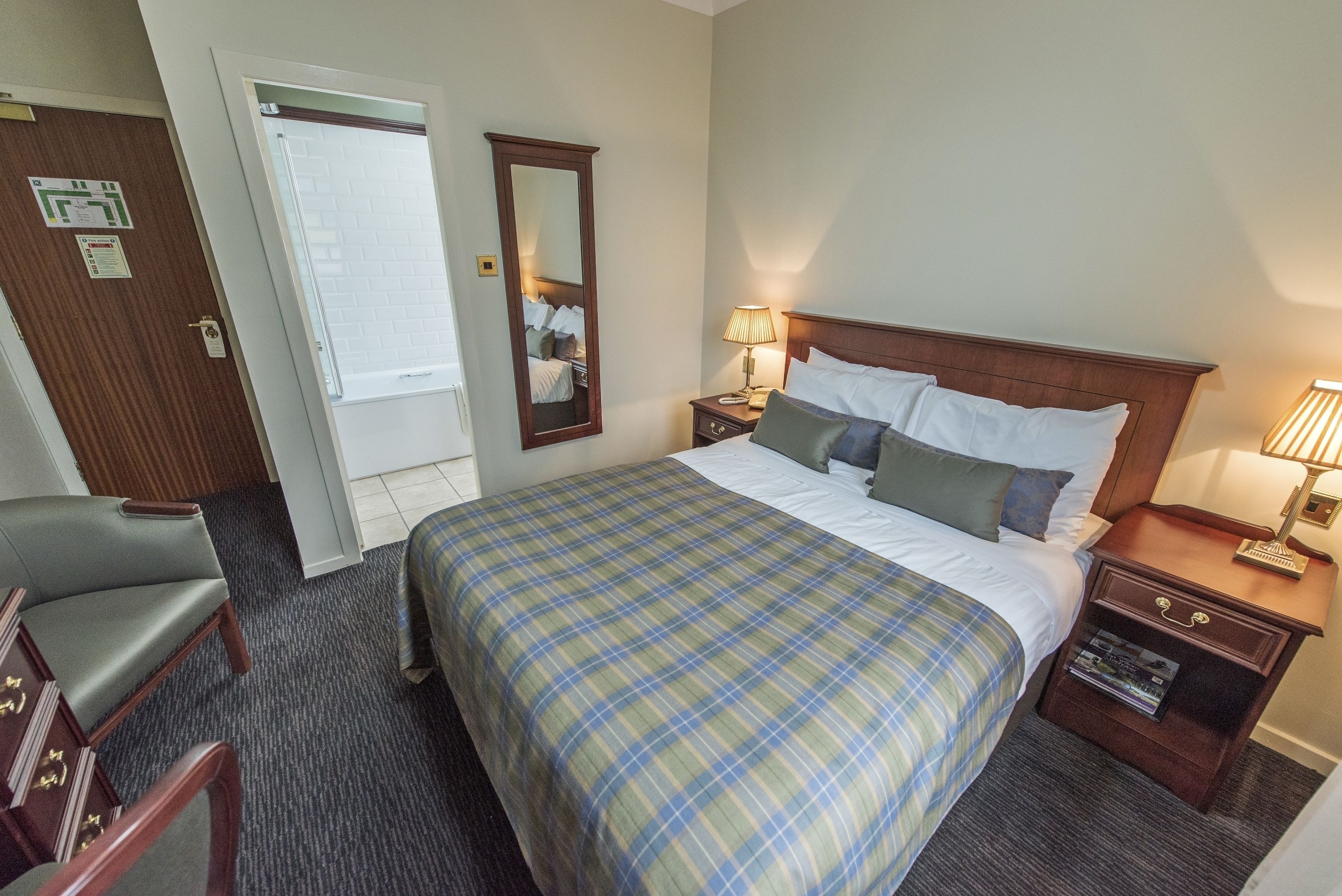 Una habitación de hotel con una cama grande, una mesita de noche con lámpara, una silla y un tocador de madera.
