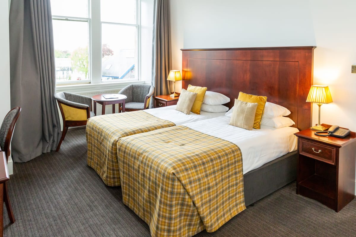 A hotel room with two beds, a desk, a chair, a window with curtains, and bedside lamps.