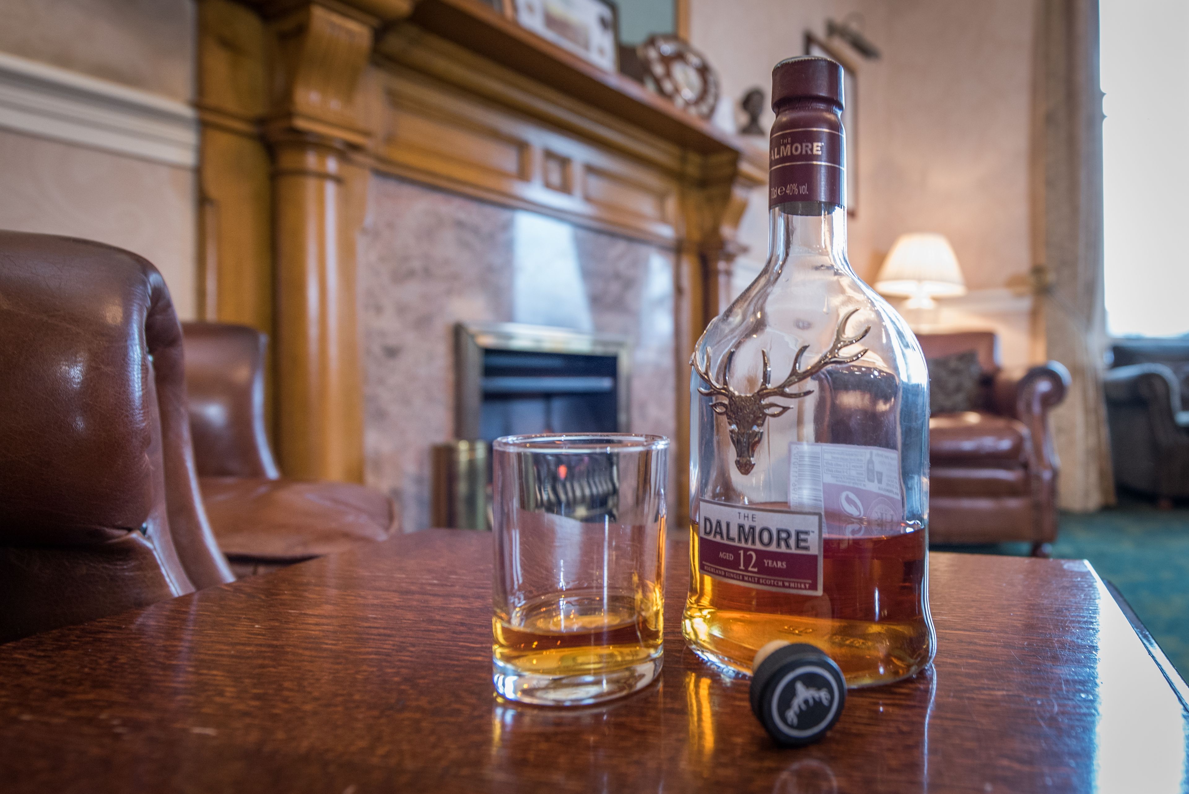 A bottle of Dalmore whiskey and a glass on a wooden table in a room with a fireplace and leather furniture.