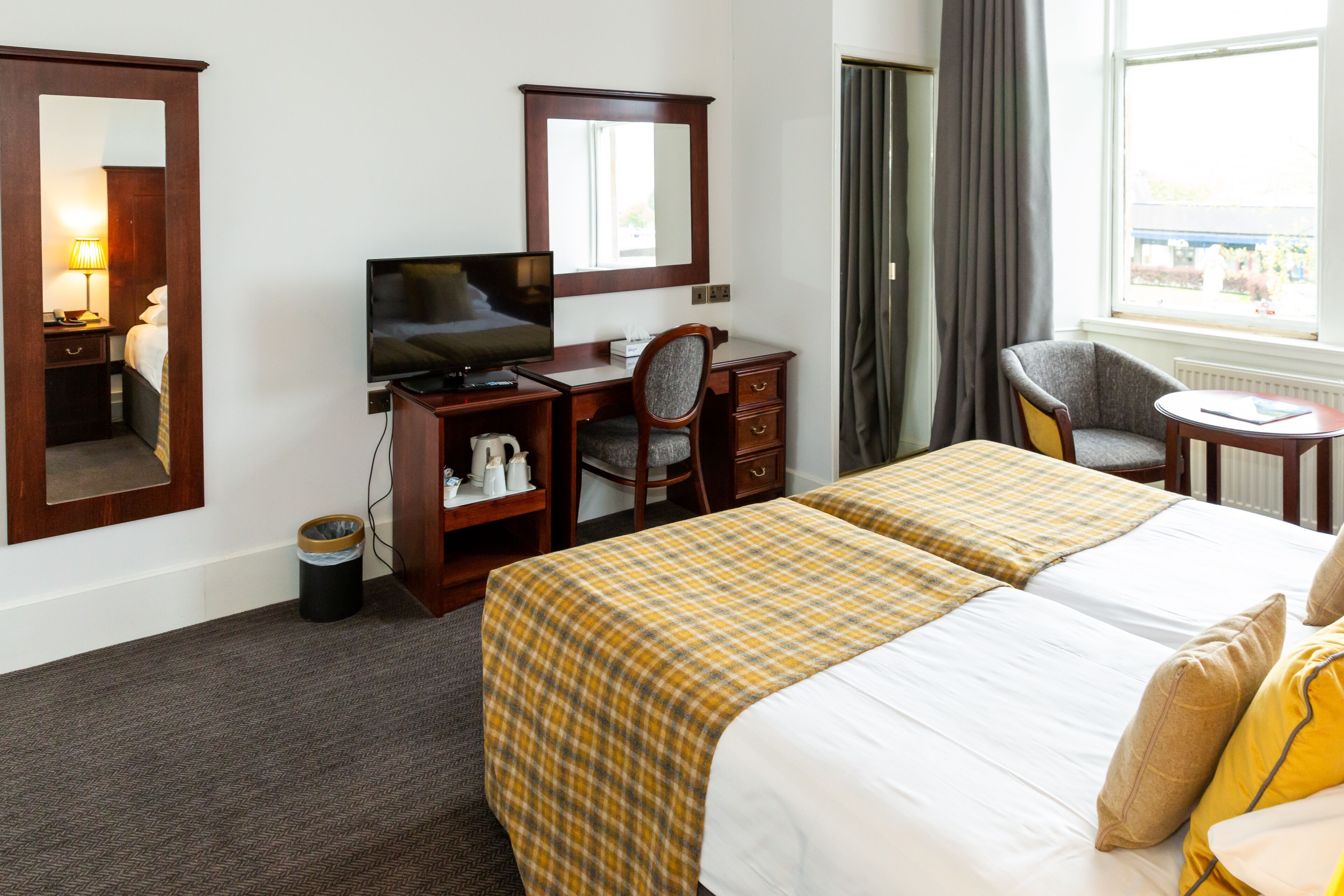 Ein Hotelzimmer mit zwei Betten, einem Schreibtisch mit Fernseher, einem Sessel, einem kleinen Tisch und einem Fenster mit Vorhängen.