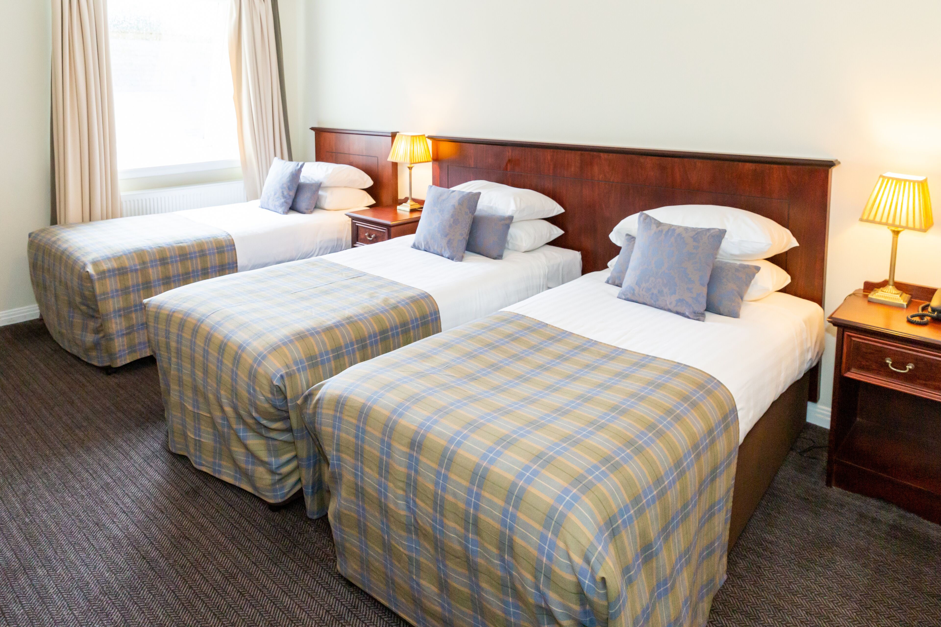 Habitación de hotel con dos camas, cada una con colcha de cuadros, cabeceros de madera, lámparas de noche y un ventanal con cortinas.