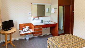 Ett hotellrum med en säng, ett litet bord med en tv och en fjärrkontroll, ett badrum med handfat och spegel samt en papperskorg.