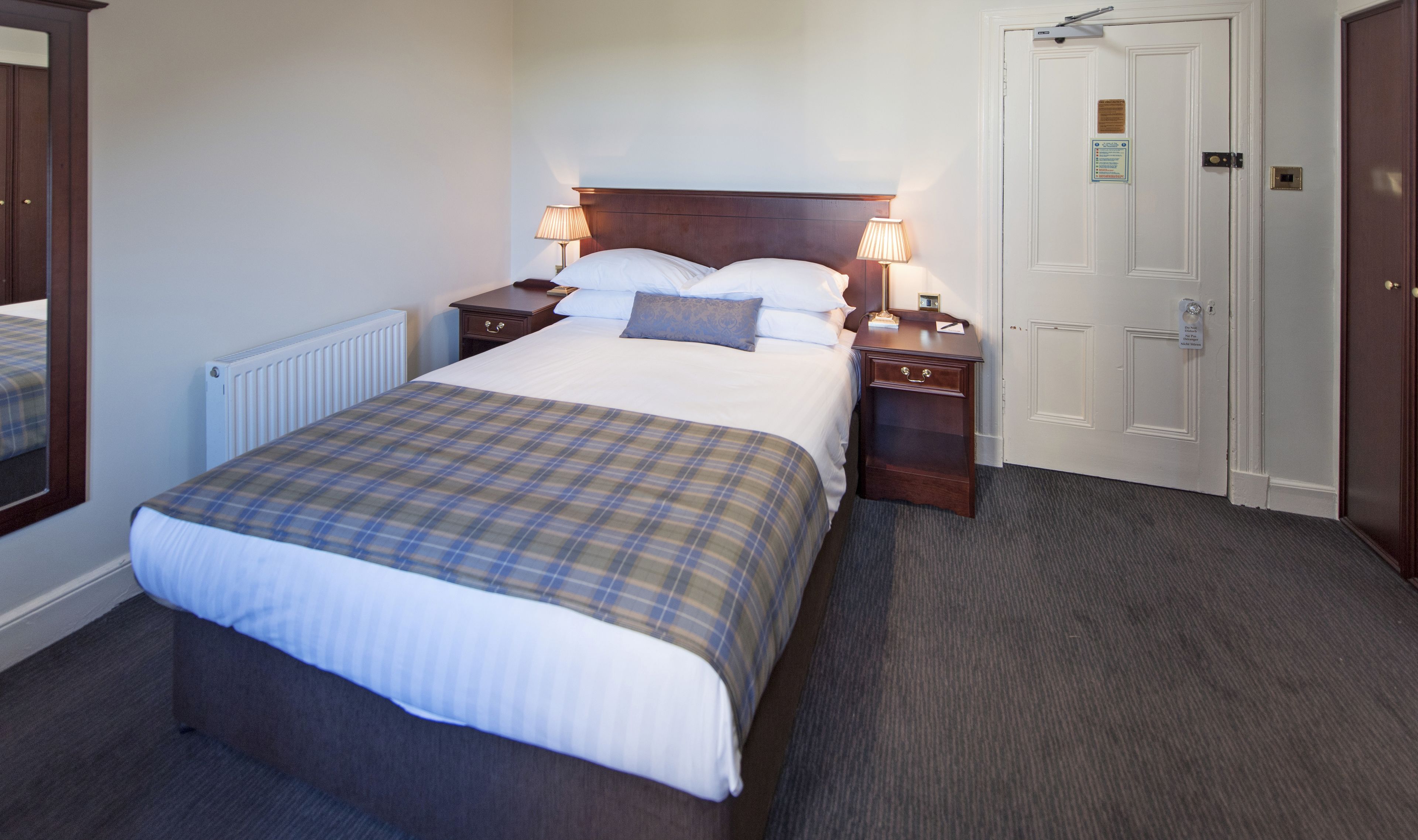 A hotel room with a large bed, two bedside tables with lamps, a wooden headboard, a white door, and a carpeted floor.
