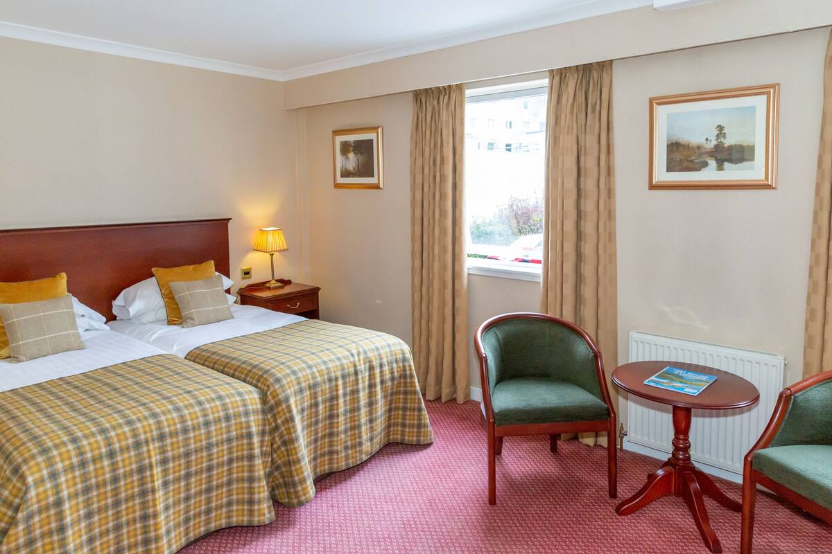 A hotel room with two beds, a green armchair, a wooden table, and a window with curtains.