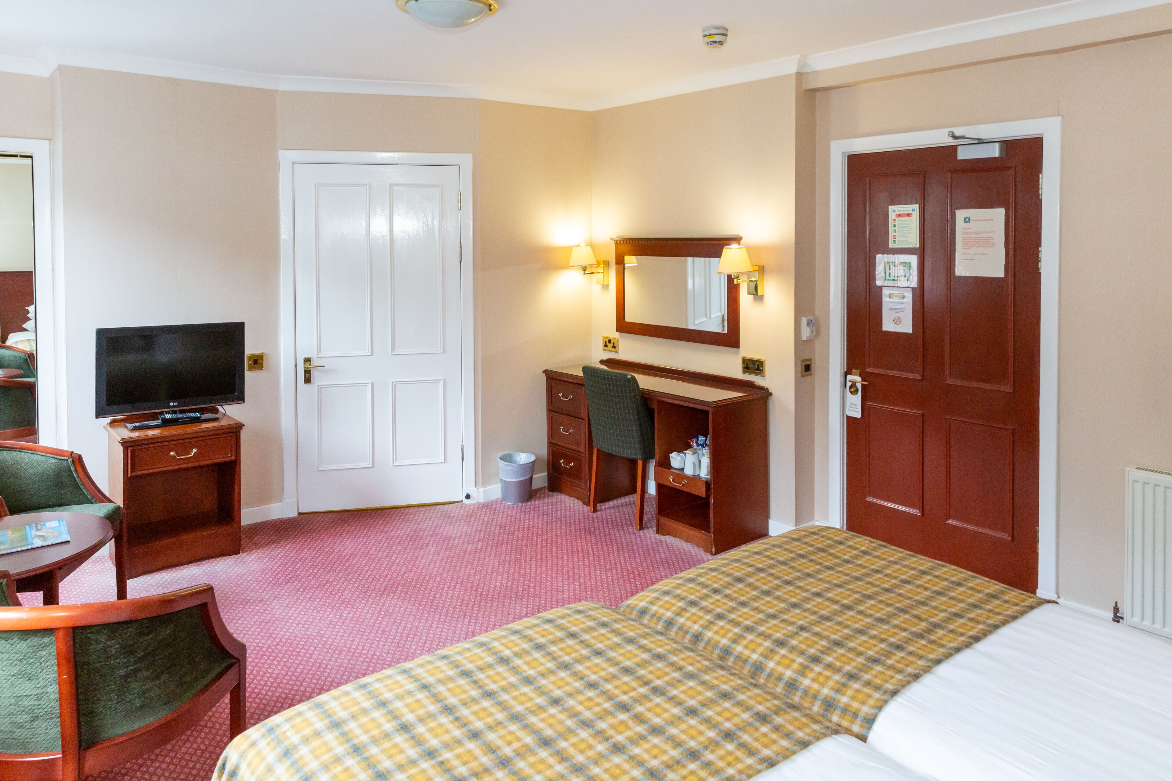 A hotel room with a bed, a desk with a chair, a television, a dresser, and a door.