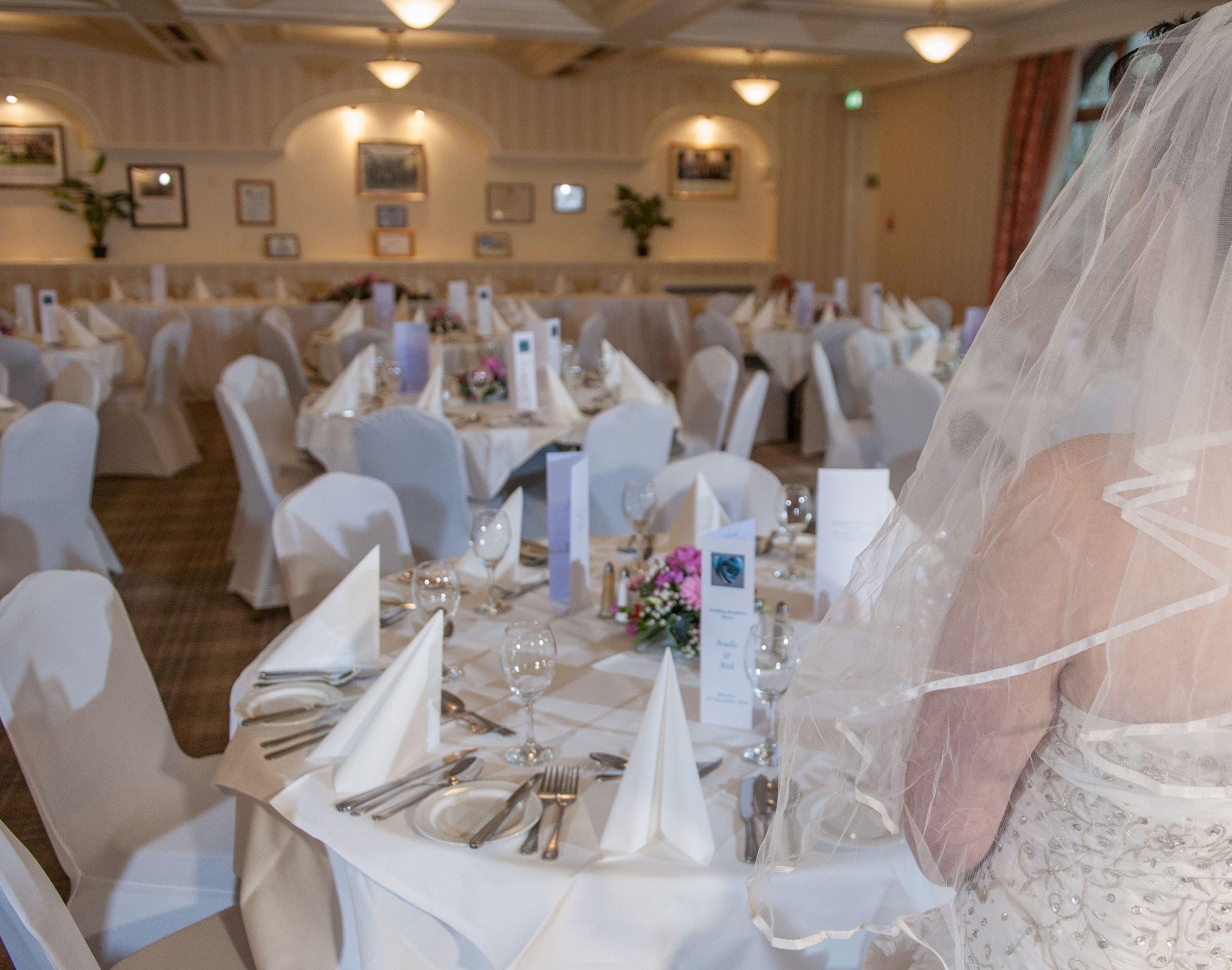 A banquet hall with round tables set for a wedding reception, featuring white tablecloths, chairs, and a bride in a white wedding dress.