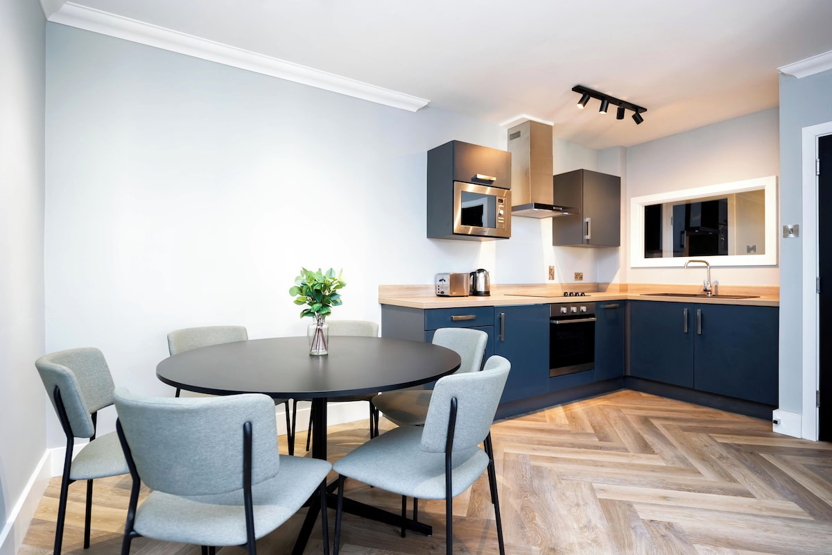 A modern kitchen with a round dining table and grey chairs, a built-in oven, and a microwave above the counter.