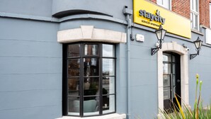 Un ventanal de esquina con marco blanco de piedra, un letrero amarillo que dice 'Staycity' arriba y una puerta pequeña con marco negro.