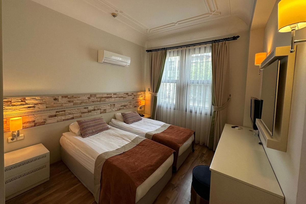 A hotel room with two beds, a desk, a television, and an air conditioning unit.