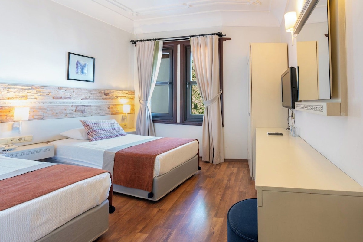 A hotel room with two beds, a desk, a TV, and a window with curtains.